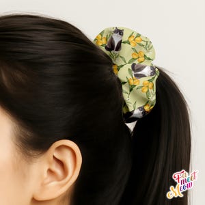 Op de afbeelding: Een lichtgroene scrunchie met een patroon van zwart-witte katten en gele citroenen. De scrunchie is om een paardenstaart van donker haar gebonden. De scrunchie heeft een bloemenontwerp met de tekst "Fa meet Meow".