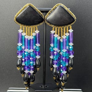 May include: A pair of vintage statement earrings. Each earring features a black triangular top with gold trim, from which hang strands of blue, purple, and clear beads, and black teardrop-shaped beads. The earrings are displayed on a black stand with a black and white striped base.