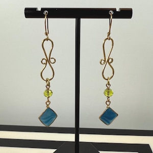 May include: A pair of gold-colored dangle earrings. Each earring features a decorative swirl at the top, a small green bead, a blue square-shaped pendant, and a gold-colored hook. The earrings are displayed on a black stand.