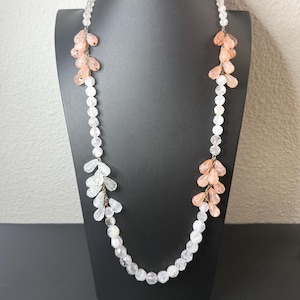 Puede incluir: Un collar largo de cuentas transparentes con racimos de acentos de cristal melocot&oacute;n y transparente. El collar se exhibe en un maniqu&iacute; negro, mostrando su delicado dise&ntilde;o y potencial para un uso elegante.