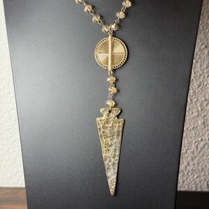 May include: A gold necklace with a crystal bead chain and a pendant featuring a round disc with a cross design and a long, pointed arrow shape.