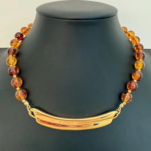 May include: A necklace featuring a series of round, translucent amber-colored beads interspersed with small gold-toned spacers. The centerpiece is a smooth, curved, polished gold-toned metal bar that connects to the beaded strands on both sides.