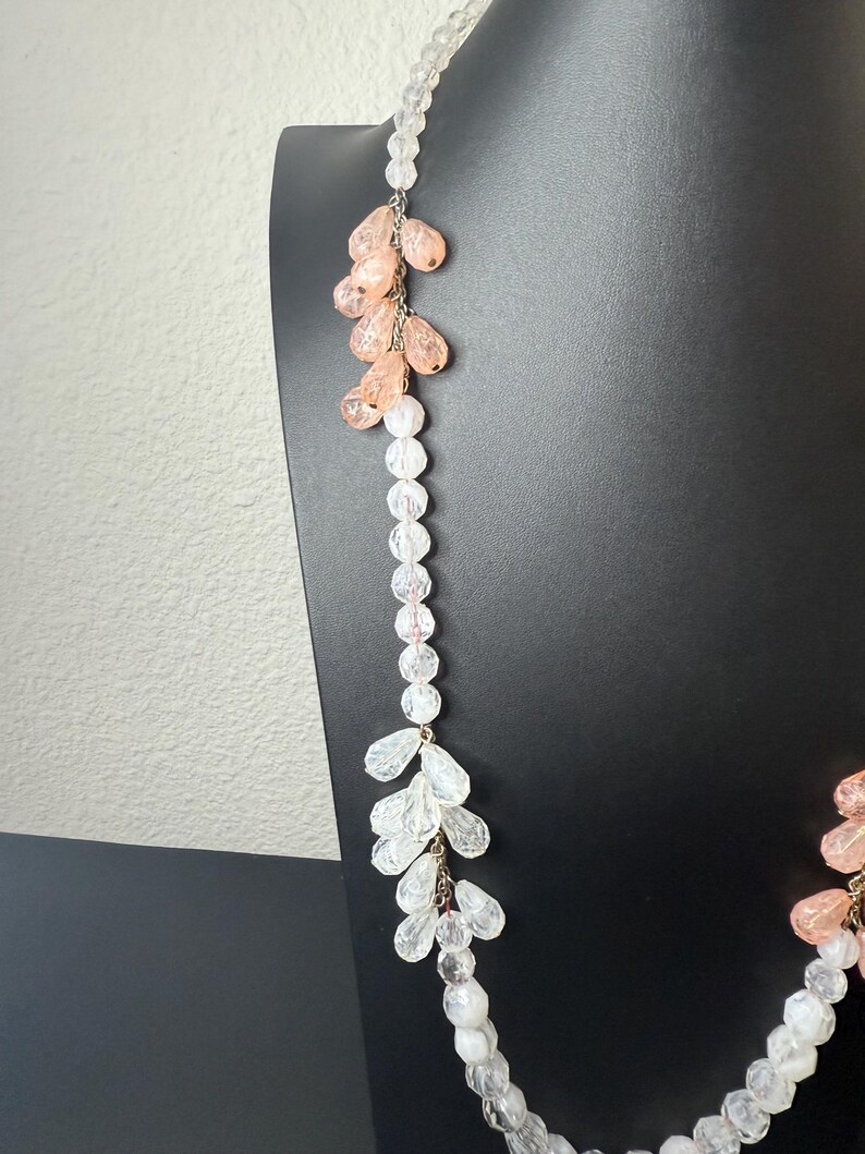 Puede incluir: Un collar largo con cuentas transparentes y color melocot&oacute;n. El collar tiene un dise&ntilde;o en cascada con racimos de cuentas en forma de l&aacute;grima de color melocot&oacute;n y cuentas claras y facetadas. El collar se exhibe en un maniqu&iacute; negro.