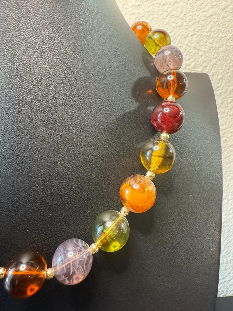 May include: A colorful beaded necklace featuring large, round beads in shades of orange, yellow, red, and purple. The beads are strung together with gold-colored connectors, creating a vibrant and eye-catching accessory.