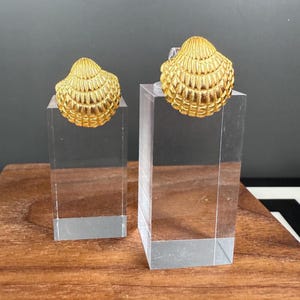 May include: Two gold-colored seashell earrings displayed on clear acrylic stands. The earrings have a detailed, textured surface, and are positioned on a wooden surface against a dark background. The earrings are a bright, shiny gold color.