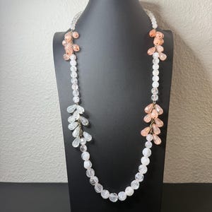 Puede incluir: Un collar largo con cuentas transparentes y color melocot&oacute;n. El collar se exhibe en un maniqu&iacute; negro. Las cuentas est&aacute;n dispuestas en un patr&oacute;n en cascada, con racimos de cuentas melocot&oacute;n y cuentas claras y facetadas.