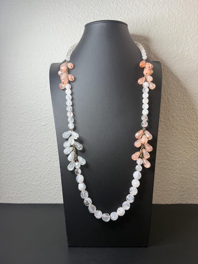 Puede incluir: Un collar largo con cuentas transparentes y color melocot&oacute;n. El collar se exhibe en un maniqu&iacute; negro. Las cuentas est&aacute;n dispuestas en un patr&oacute;n en cascada, con racimos de cuentas melocot&oacute;n y cuentas claras y facetadas.