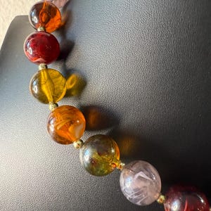 May include: A colorful beaded necklace with large, round beads in shades of red, orange, yellow, green, and purple. The beads are strung together with small gold-colored connectors, creating a vibrant and eye-catching accessory.