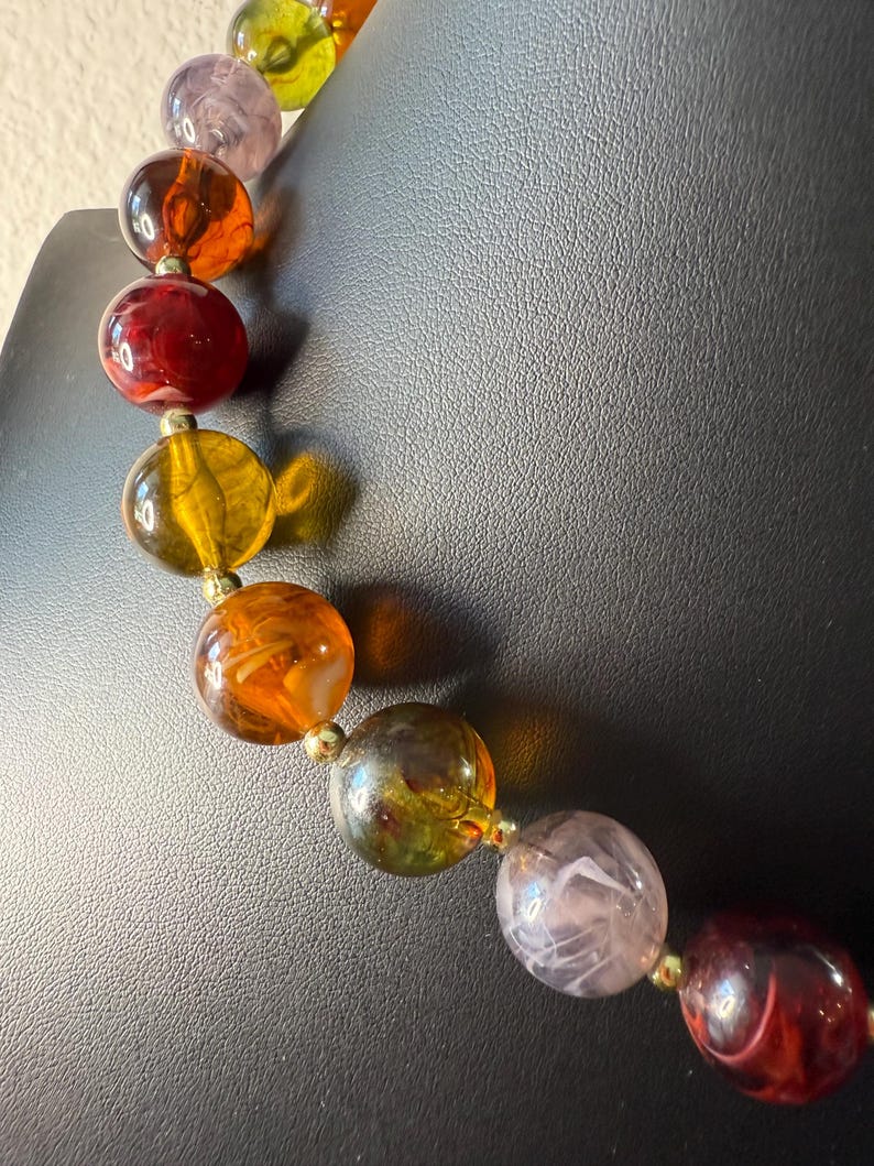 May include: A colorful beaded necklace with large, round beads in shades of red, orange, yellow, green, and purple. The beads are strung together with small gold-colored connectors, creating a vibrant and eye-catching accessory.