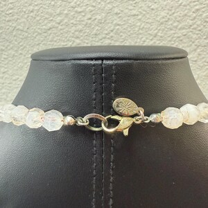 Puede incluir: Primer plano de un collar de cuentas transparentes y facetadas con un cierre plateado y un colgante dorado. Las cuentas est&aacute;n ensartadas, creando un accesorio delicado y elegante. El collar se exhibe en un maniqu&iacute; negro.