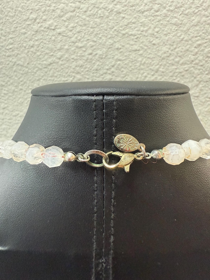 Puede incluir: Primer plano de un collar de cuentas transparentes y facetadas con un cierre plateado y un colgante dorado. Las cuentas est&aacute;n ensartadas, creando un accesorio delicado y elegante. El collar se exhibe en un maniqu&iacute; negro.