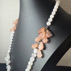 Puede incluir: Un collar largo con cuentas transparentes y color melocot&oacute;n. El collar se exhibe en un maniqu&iacute; negro. Las cuentas est&aacute;n ensartadas con una cadena plateada. Las cuentas color melocot&oacute;n tienen forma de l&aacute;grima y est&aacute;n agrupadas.