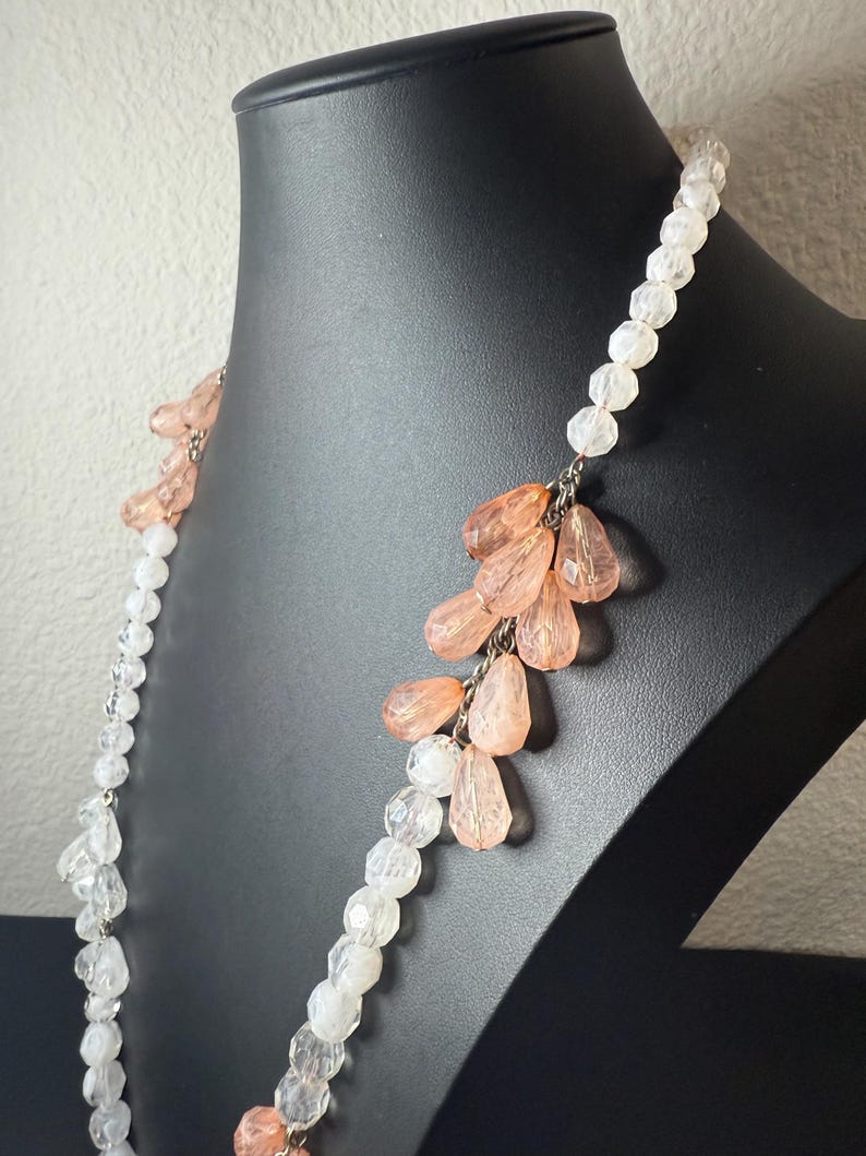 Puede incluir: Un collar largo con cuentas transparentes y color melocot&oacute;n. El collar se exhibe en un maniqu&iacute; negro. Las cuentas est&aacute;n ensartadas con una cadena plateada. Las cuentas color melocot&oacute;n tienen forma de l&aacute;grima y est&aacute;n agrupadas.