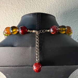 May include: A beaded necklace with large, round beads in shades of amber, red, and green. The necklace is displayed on a black mannequin. The clasp is silver-toned, with a chain and a single red bead at the end.