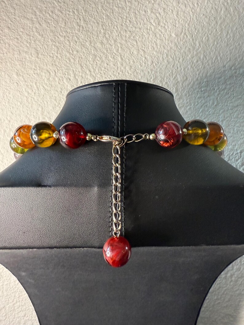 May include: A beaded necklace with large, round beads in shades of amber, red, and green. The necklace is displayed on a black mannequin. The clasp is silver-toned, with a chain and a single red bead at the end.