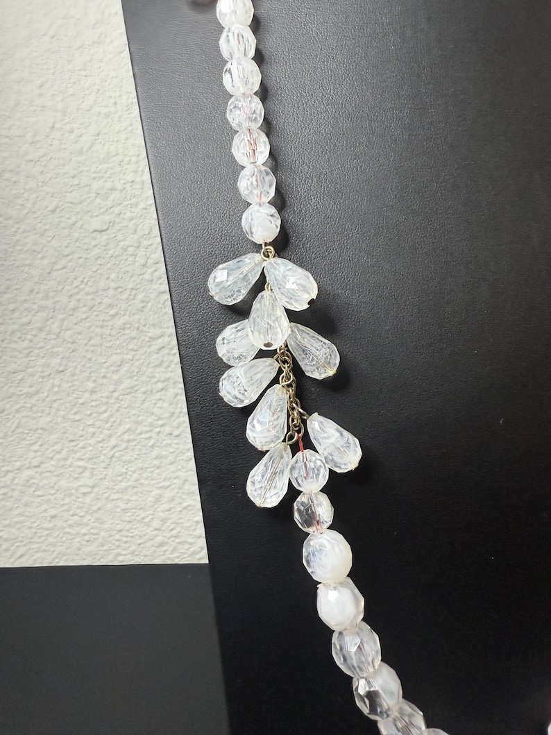 Puede incluir: Un collar de cuentas transparentes con un dise&ntilde;o en cascada. El collar presenta una hebra de cuentas redondas y un grupo de cuentas en forma de l&aacute;grima. Las cuentas est&aacute;n conectadas con metal dorado. El collar se muestra sobre un fondo negro.