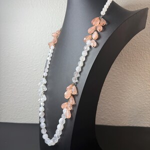 Puede incluir: Un collar largo con cuentas transparentes y color melocot&oacute;n. El collar se exhibe en un maniqu&iacute; negro. Las cuentas est&aacute;n dispuestas en un patr&oacute;n en cascada, con las cuentas color melocot&oacute;n agrupadas. El collar es una joya delicada.