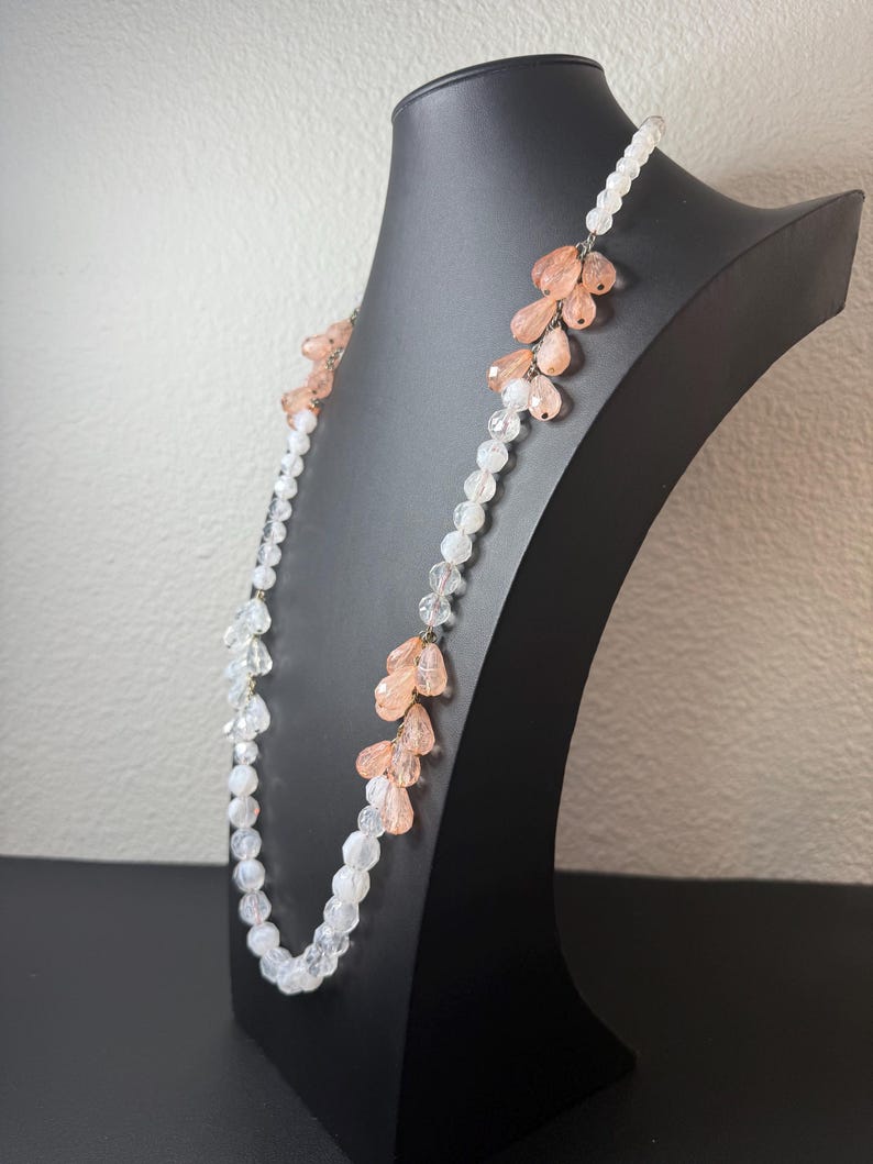 Puede incluir: Un collar largo con cuentas transparentes y color melocot&oacute;n. El collar se exhibe en un maniqu&iacute; negro. Las cuentas est&aacute;n dispuestas en un patr&oacute;n en cascada, con las cuentas color melocot&oacute;n agrupadas. El collar es una joya delicada.