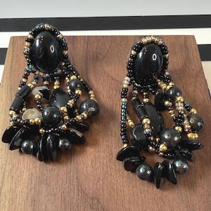May include: A pair of black beaded earrings with a large oval bead at the top. The earrings feature a mix of black, gold, and iridescent beads, with dangling accents. The earrings are displayed on a wooden surface.