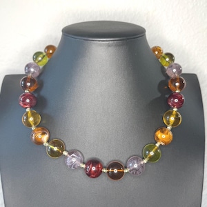 May include: A colorful beaded necklace displayed on a black mannequin. The necklace features large, round beads in shades of yellow, orange, purple, green, and burgundy. The beads are strung together with silver-colored metal.