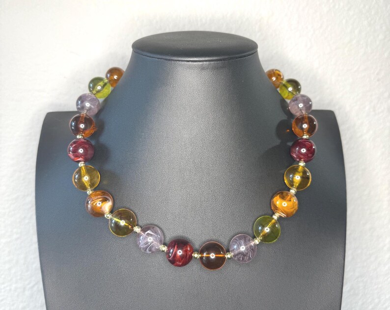 May include: A colorful beaded necklace displayed on a black mannequin. The necklace features large, round beads in shades of yellow, orange, purple, green, and burgundy. The beads are strung together with silver-colored metal.