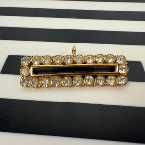 May include: A rectangular gold-toned brooch with a central open space, surrounded by two rows of clear, round gemstones. The brooch has a small loop at the top for attachment. The background is a black and white striped pattern.