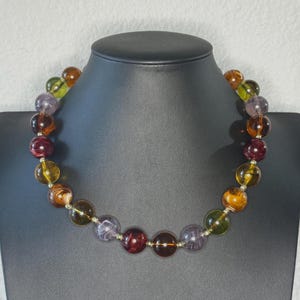 May include: A beaded necklace with large, round beads in shades of amber, purple, green, and burgundy. The beads are strung on a silver-colored chain and displayed on a black jewelry bust. The necklace is a statement piece.