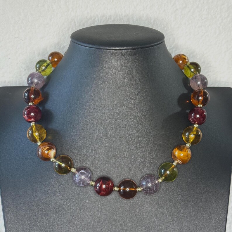 May include: A beaded necklace with large, round beads in shades of amber, purple, green, and burgundy. The beads are strung on a silver-colored chain and displayed on a black jewelry bust. The necklace is a statement piece.