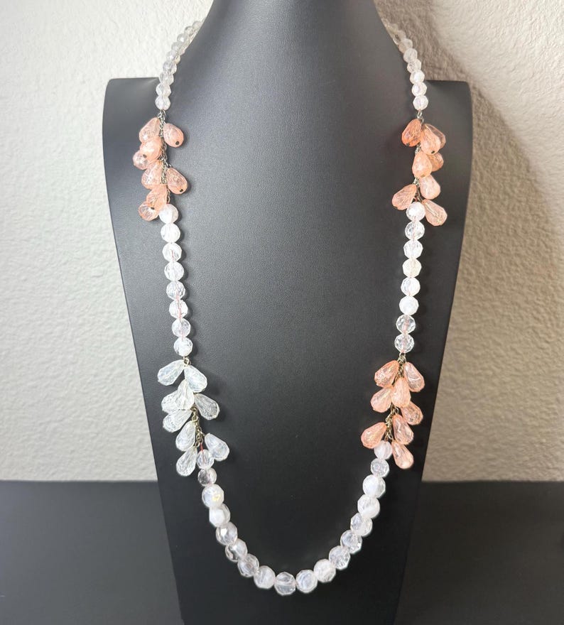 Puede incluir: Un collar largo de cuentas transparentes con racimos de acentos de cristal melocot&oacute;n y transparente. El collar se exhibe en un maniqu&iacute; negro, mostrando su delicado dise&ntilde;o y potencial para un uso elegante.