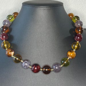 May include: A colorful beaded necklace featuring large, round beads in shades of amber, purple, green, and burgundy. The beads are strung on a delicate silver-toned chain, creating a striking contrast. The necklace is displayed on a black mannequin.