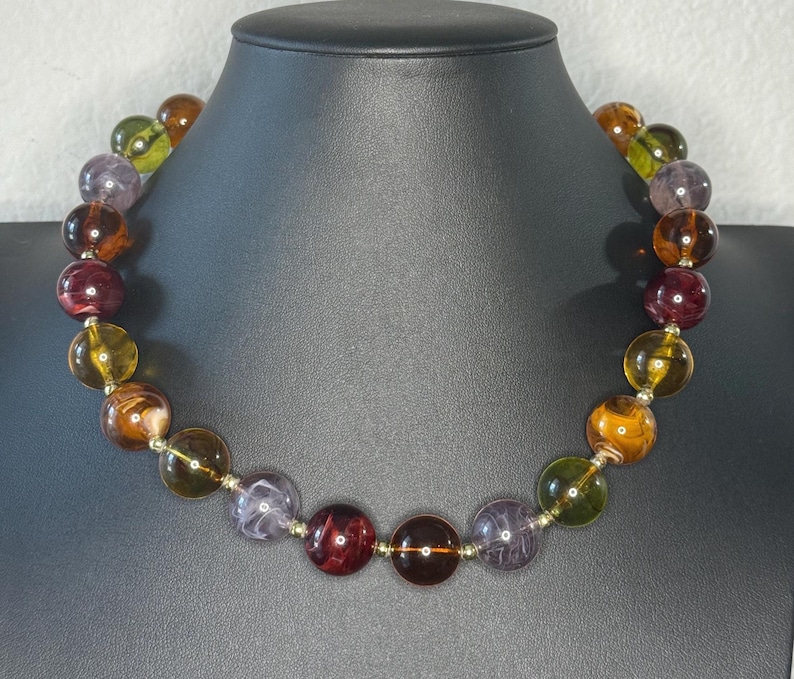 May include: A colorful beaded necklace featuring large, round beads in shades of amber, purple, green, and burgundy. The beads are strung on a delicate silver-toned chain, creating a striking contrast. The necklace is displayed on a black mannequin.