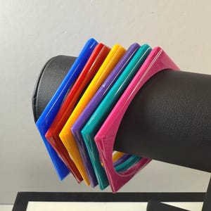 May include: A collection of colorful square bangles in shades of blue, red, yellow, purple, teal, and pink. The bangles are displayed on a black stand, showcasing their vibrant colors and geometric design. These bold accessories add a pop of color to any outfit.