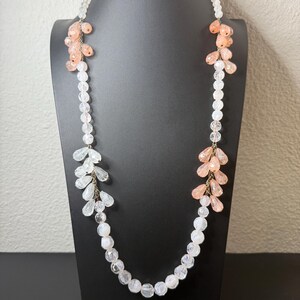 Puede incluir: Un collar largo con cuentas transparentes y color melocot&oacute;n. El collar presenta una hebra de cuentas redondas transparentes con racimos de cuentas en forma de l&aacute;grima de color melocot&oacute;n y transparentes. El collar se exhibe en un maniqu&iacute; negro.
