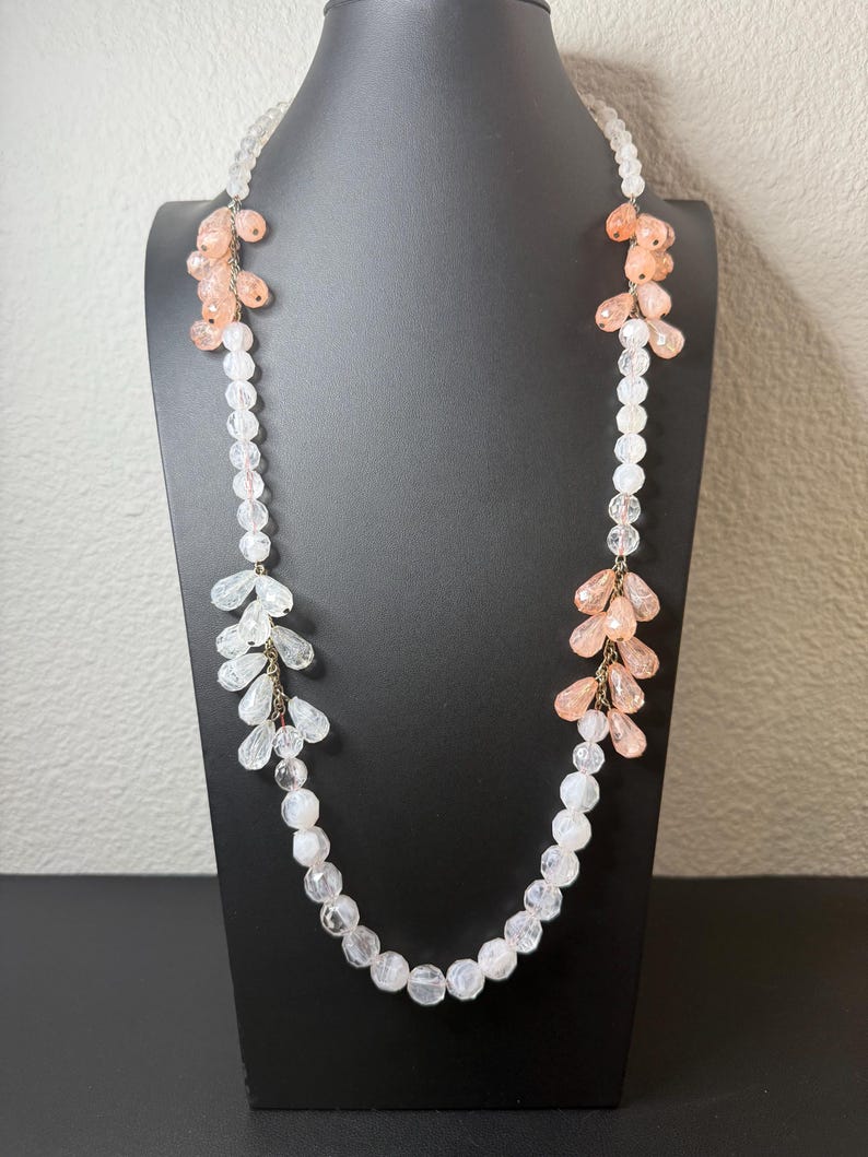 Puede incluir: Un collar largo con cuentas transparentes y color melocot&oacute;n. El collar presenta una hebra de cuentas redondas transparentes con racimos de cuentas en forma de l&aacute;grima de color melocot&oacute;n y transparentes. El collar se exhibe en un maniqu&iacute; negro.