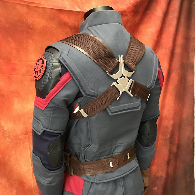 May include: A blue tactical jacket featuring red accents, brown shoulder straps with a silver metallic harness piece, and a circular black and red Hydra emblem on the right shoulder. The garment includes dark blue elbow patches and a brown utility belt.