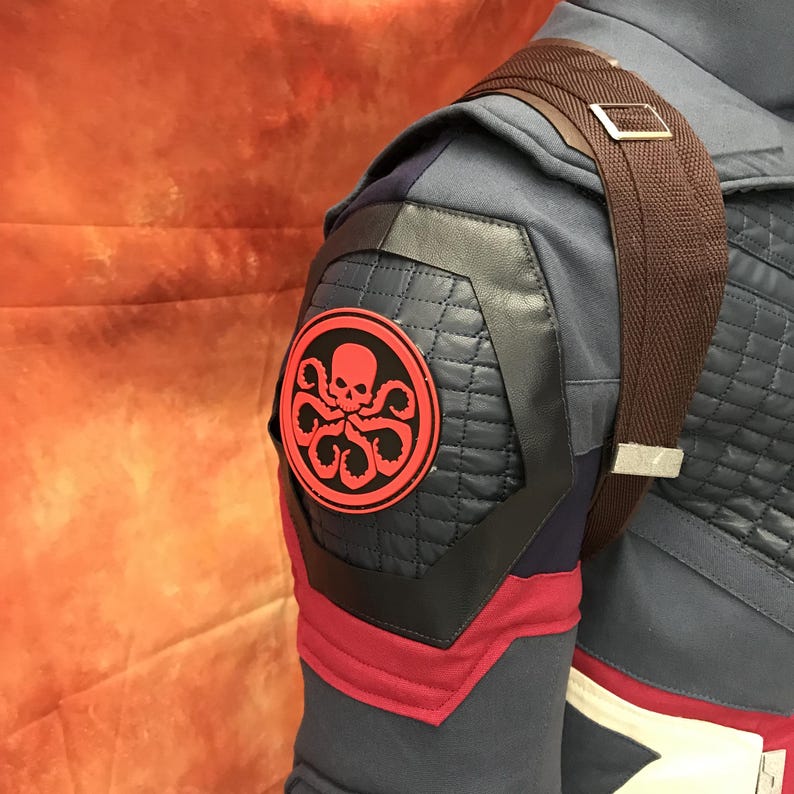 May include: A dark blue tactical jacket featuring a quilted shoulder panel with a circular red and black Hydra emblem. The garment includes a brown fabric strap with a silver metal buckle, dark blue fabric sections, and red trim accents.