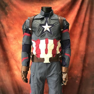 May include: A Captain America costume featuring a dark blue tactical jacket with a white star emblem on the chest, red and white vertical stripes on the midsection, brown leather-like straps, a utility belt, and matching dark blue trousers.