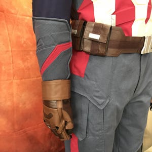 May include: A costume ensemble featuring blue and red fabric pants and a matching long-sleeved top with a red chest panel. A brown utility belt with a rectangular pouch and a brown leather-like glove with a matching forearm gauntlet are worn.