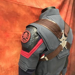 May include: A blue tactical jacket featuring a red and black circular Hydra emblem on the shoulder, quilted black shoulder panels, red fabric accents, and a brown harness system with a silver metallic center piece worn over the shoulders.