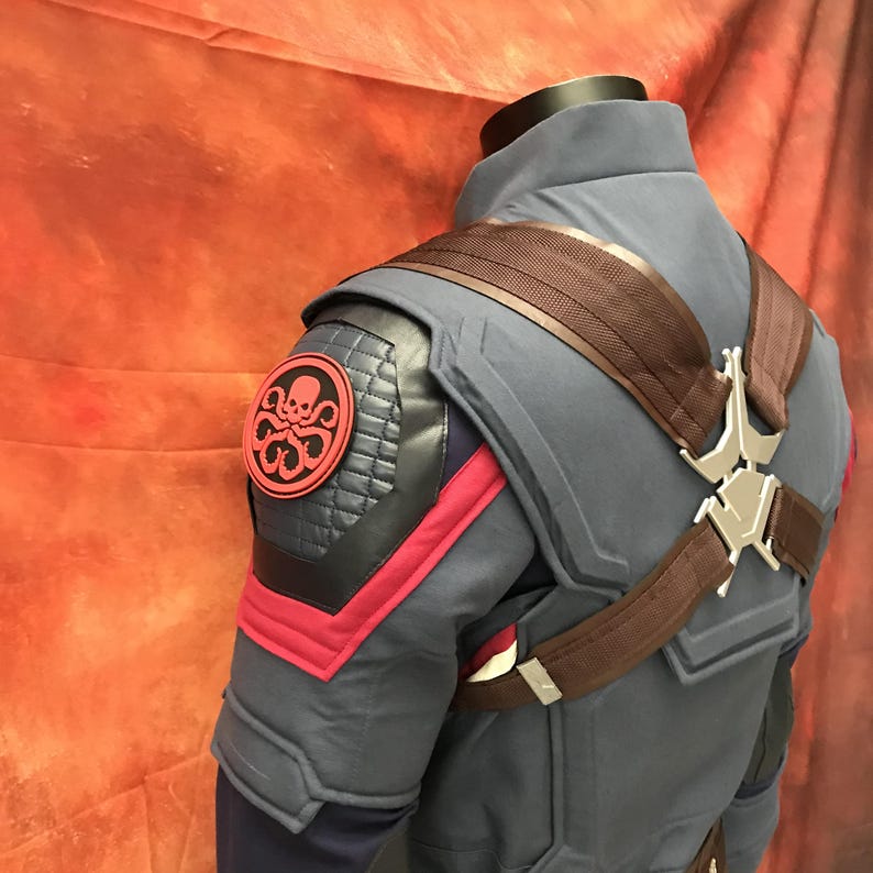 May include: A blue tactical jacket featuring a red and black circular Hydra emblem on the shoulder, quilted black shoulder panels, red fabric accents, and a brown harness system with a silver metallic center piece worn over the shoulders.