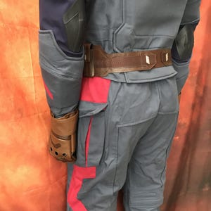May include: A tactical costume set featuring a grey jacket with dark blue sleeves and black elbow pads, paired with matching grey trousers with red side panels. A brown utility belt with a pouch and metallic accents is worn around the waist.