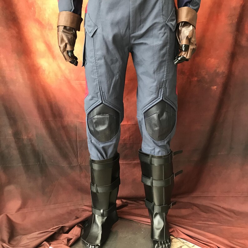 May include: A blue jumpsuit featuring dark blue knee pads, brown fingerless gloves, and black calf-high boots with multiple horizontal straps. The garment includes structured seams and utility-style pockets on the upper thighs.