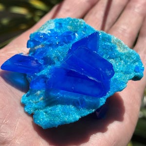 May include: A vibrant blue crystal cluster with sharp, geometric formations sits atop a turquoise matrix. The crystal's intense color contrasts with the lighter blue-green base. This mineral specimen is held in a hand, showcasing its natural beauty.