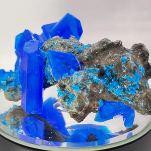 May include: A vibrant display of blue crystals and mineral formations. The crystals are a striking shade of bright blue, contrasting with the darker, textured rock base. The arrangement is presented on a reflective surface, enhancing the visual appeal.