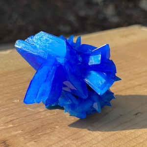 May include: A vibrant, crystalline cluster of deep blue crystals. The mineral formation has sharp, angular facets and a translucent quality, resting on a light brown wooden surface. The crystals are illuminated by natural light, highlighting their color and structure.