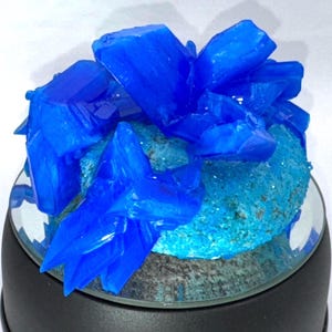 May include: A vibrant blue crystal cluster, with large, sharp formations, sits atop a textured, turquoise-colored base. The crystals are displayed on a clear, circular stand, creating a striking contrast against the white background.