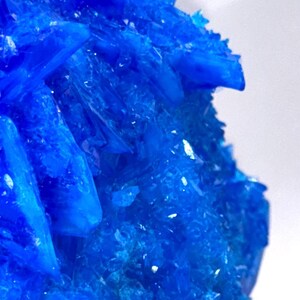 May include: Close-up of a vibrant, crystalline formation. The object is a deep, saturated blue, with sharp, angular facets and a textured surface. The crystals are clustered together, creating a visually striking display.