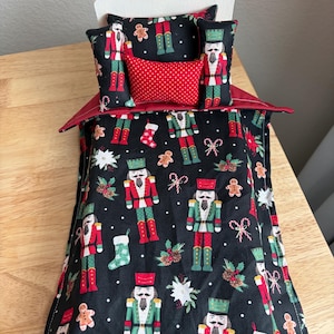 May include: A miniature wooden bed frame featuring a black fabric bedding set with a festive pattern of nutcrackers, gingerbread men, candy canes, and stockings. The set includes a quilt, two rectangular pillows, and a small red polka-dot pillow.