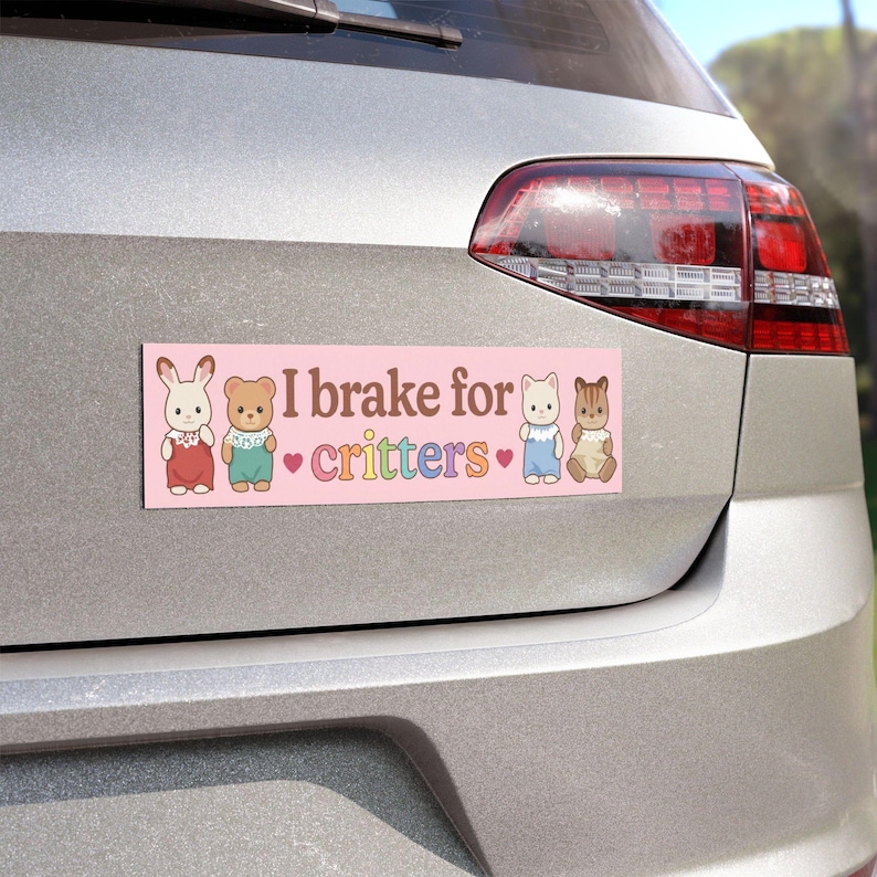 Op de afbeelding: Een roze bumpersticker met de tekst "I brake for critters" en illustraties van een konijn, beer, kat en eekhoorn. De sticker is rechthoekig en bevestigd aan de achterkant van een zilveren auto.