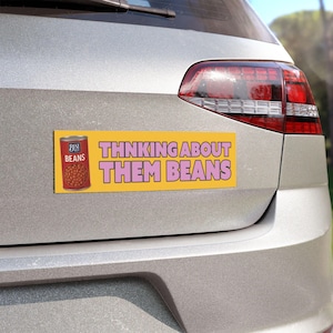 May include: A yellow bumper sticker with the text "THINKING ABOUT THEM BEANS" in pink letters. The sticker also features a can of beans with the words "BEST BEANS" on the label. The sticker is attached to the back of a silver car.